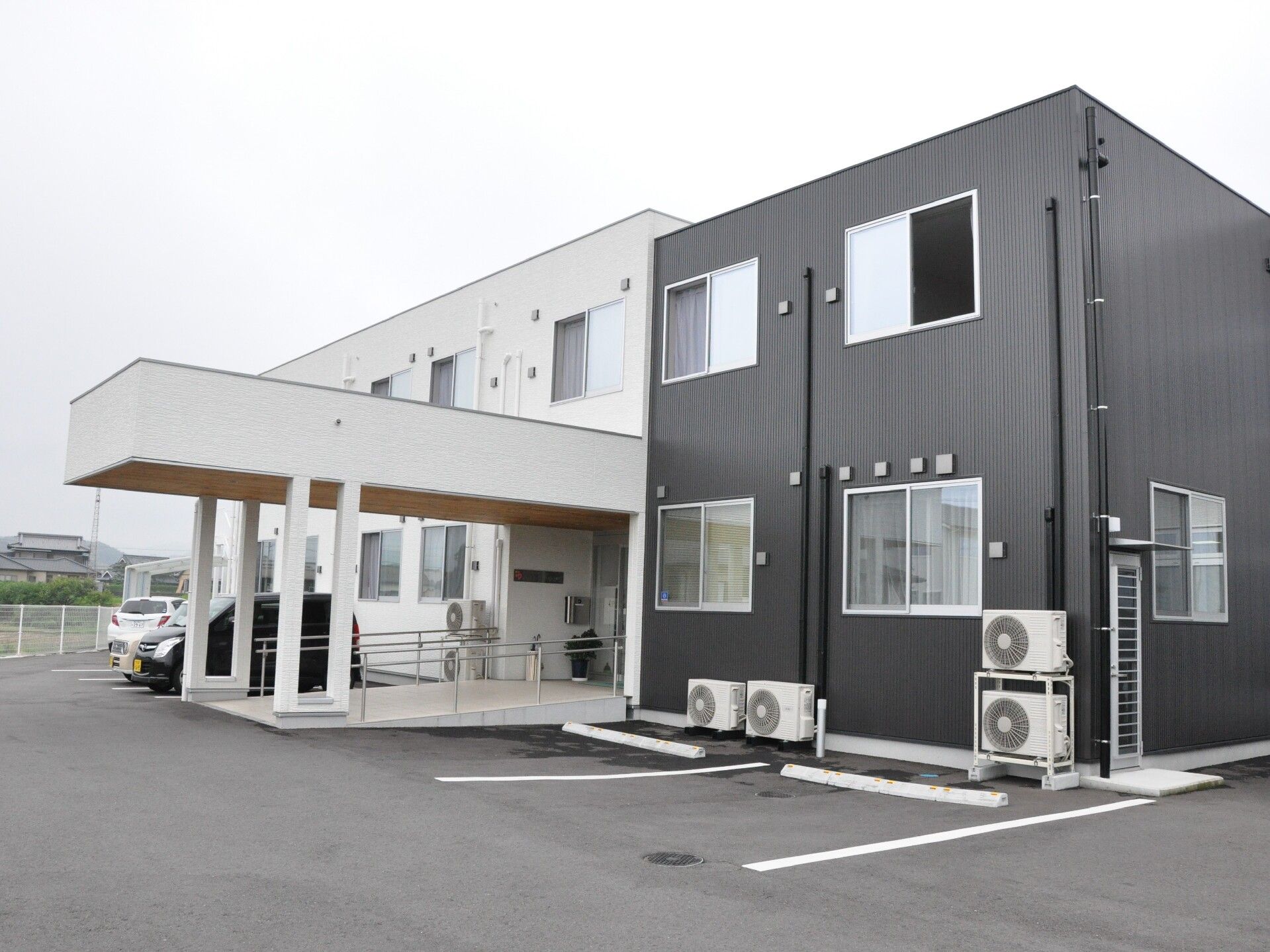白とダークグレーの2階建ての建物は玄関先に大きな車寄せが付き、手すり付きのスロープが付いている。