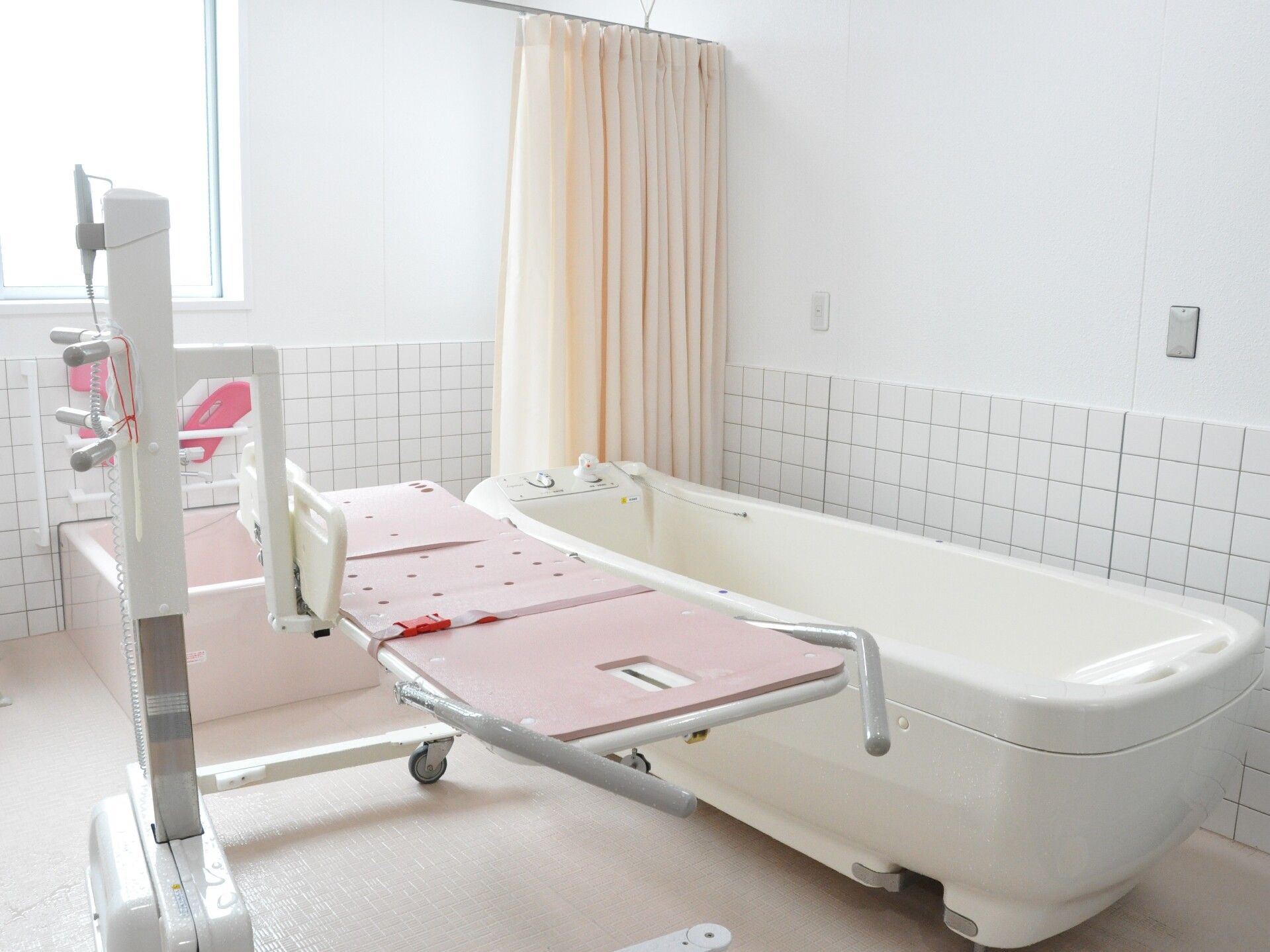 手前には白とピンクの寝台浴が設置され、奥には３方向から介助ができる一人用の浴槽があり、中央にはカーテンがある。