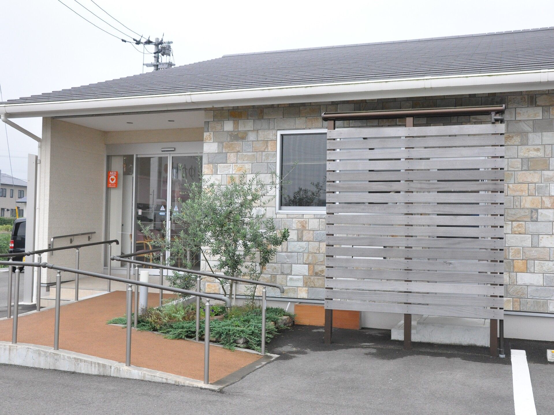 平屋建ての建物の玄関には手すり付きのスロープが付き、AEDのステッカーも貼られ、植物も植えられている。