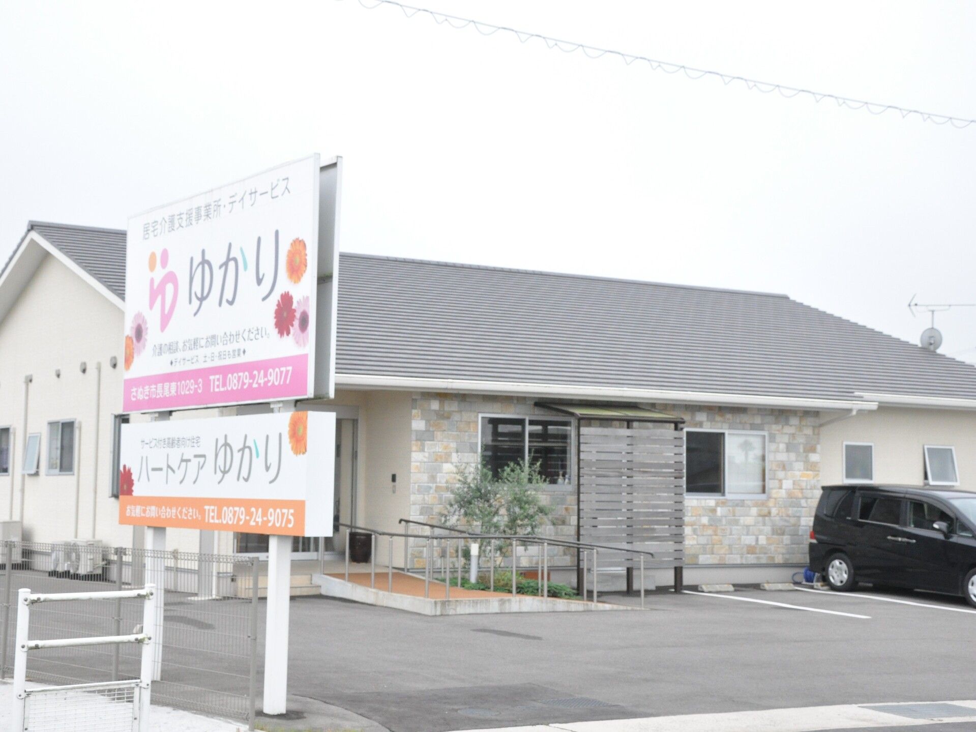 駐車場が付いた平屋建ての建物の前にはピンクとオレンジの看板が建っている。サービス付き高齢者向け住宅と併設事業所のもの。