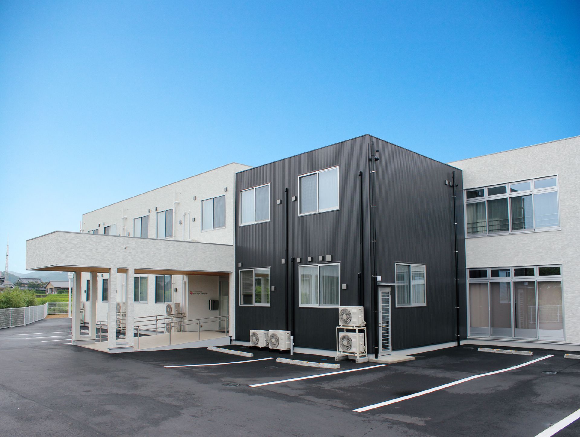 白とダークグレーの2階建ての建物は玄関に手すり付きのスロープが付いている。駐車場も広い。