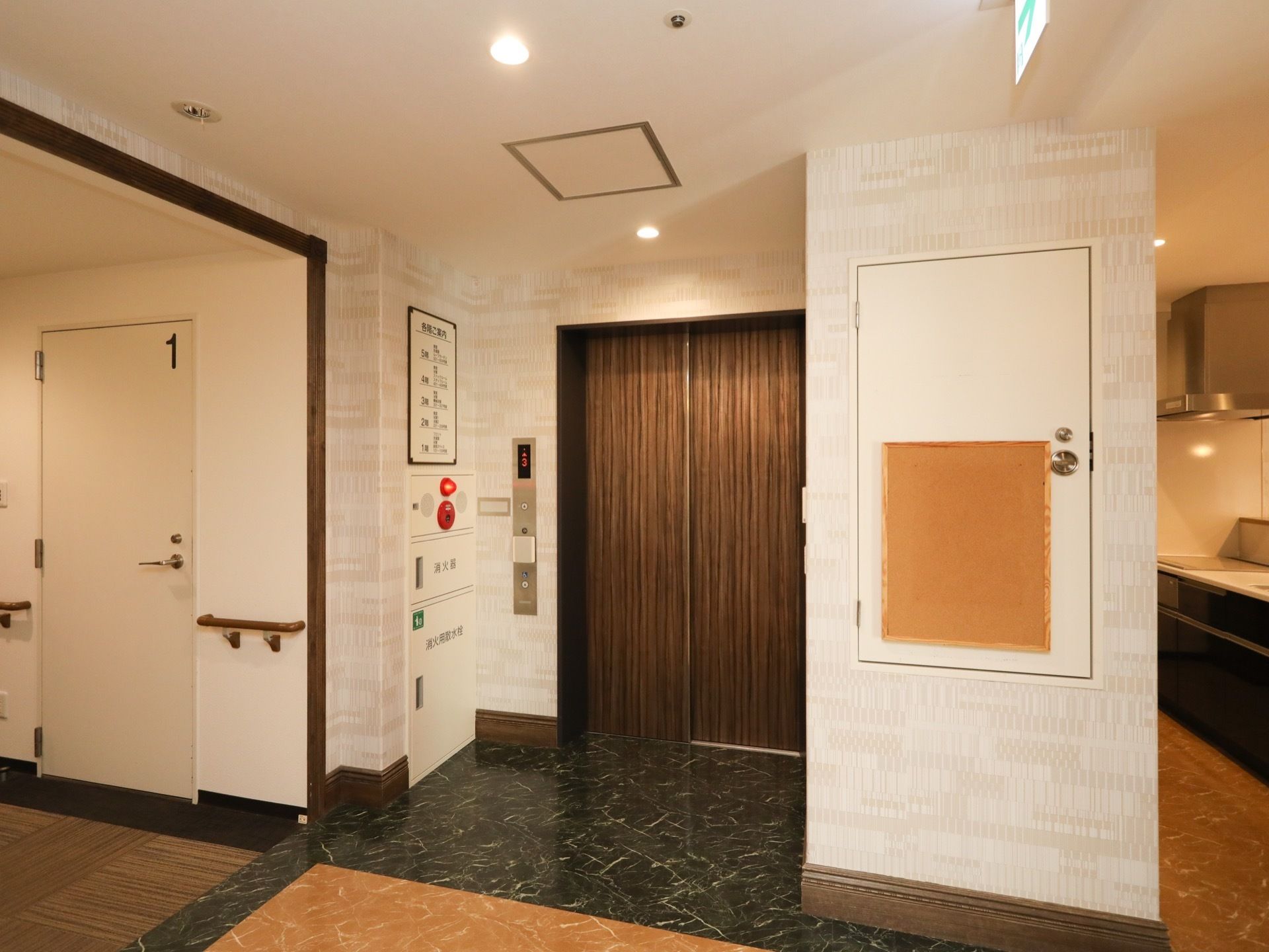 木目調の扉のエレベーターが設置されている。乗降口の左側に操作パネルがあり、その横の壁に館内案内図が表示されている。