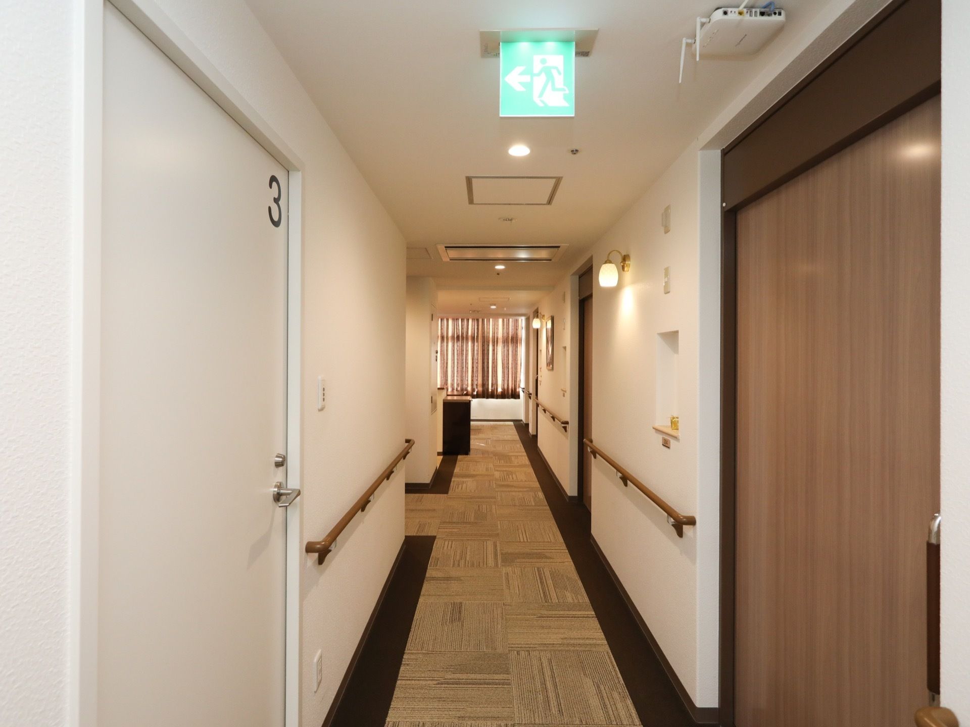 廊下の床面に、茶色を基調としたカーペットが敷かれている。天井に、避難階段の場所を示す緑色のサインが表示されている。