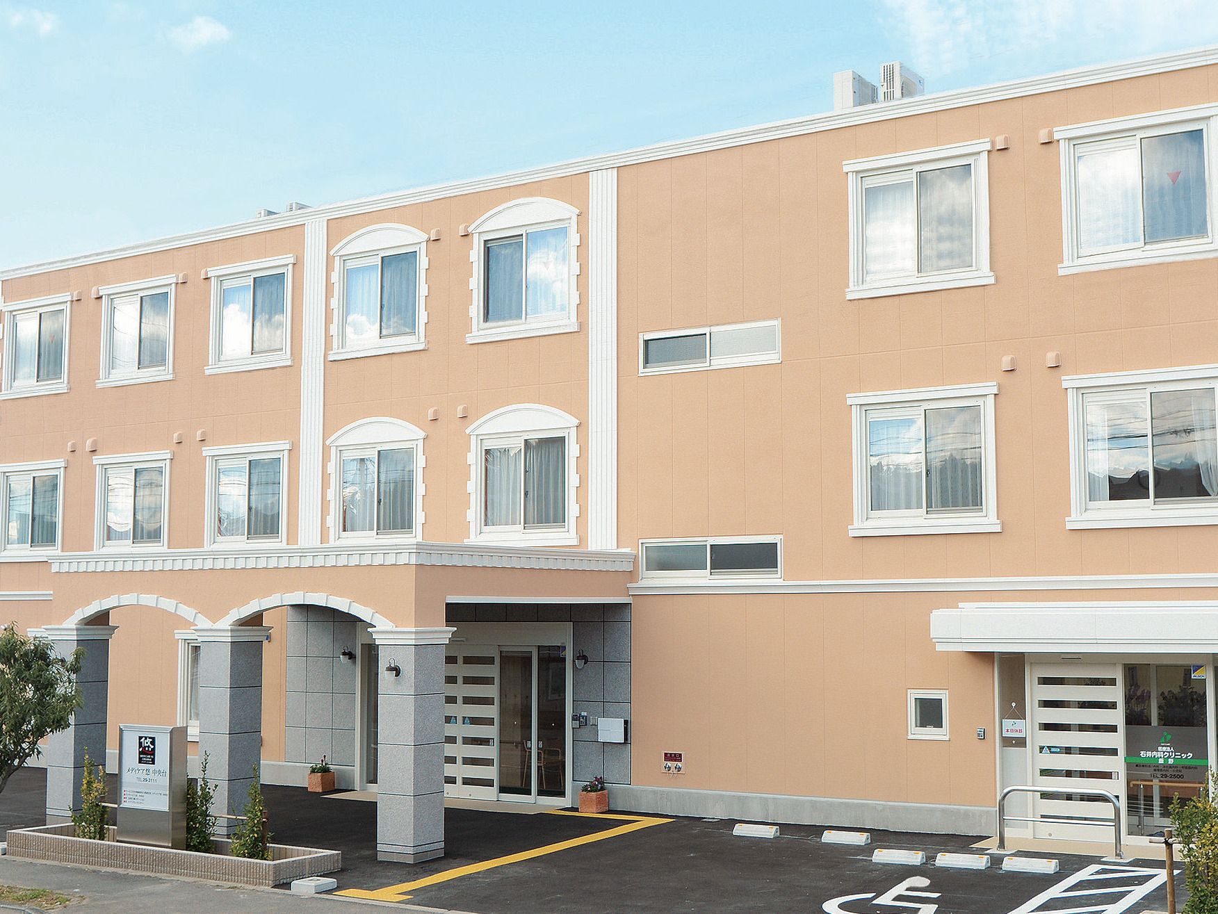 ベージュの3階建ての建物。玄関には車寄せが付き、車いすの男性を女性スタッフが介助している。