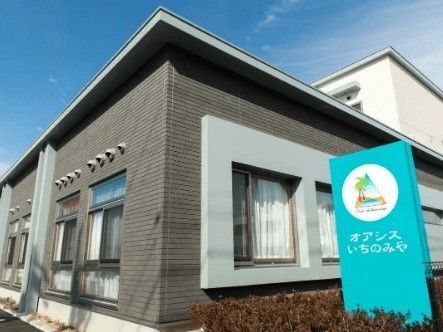 白とグレーの外壁。緑の看板にはイラストも描かれ、垣根の中に立てられている。