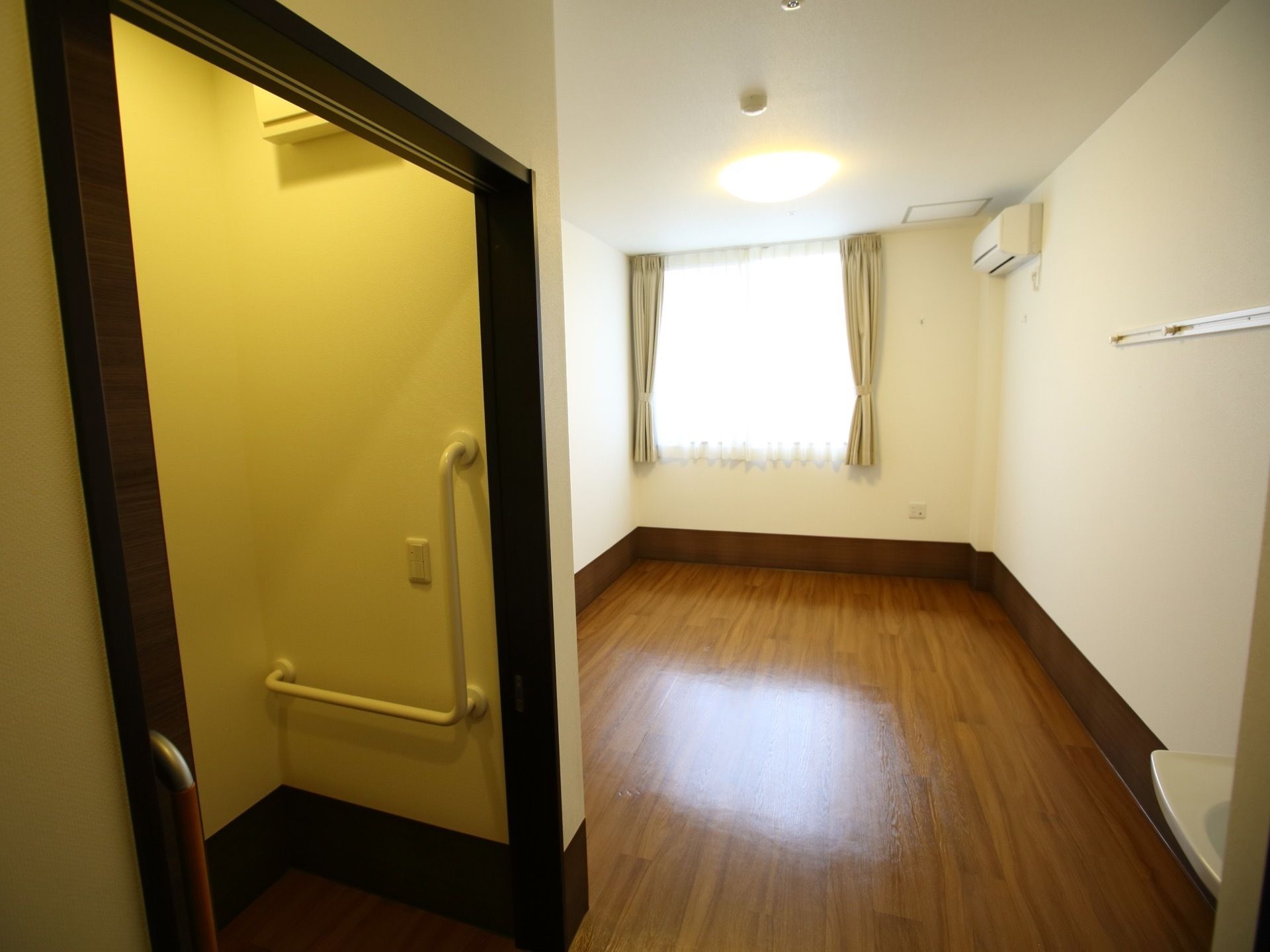 ご入居前の居室である。木目調のフローリングに白い壁が設計され、エアコンやカーテンが取り付けられた窓が設置されている。