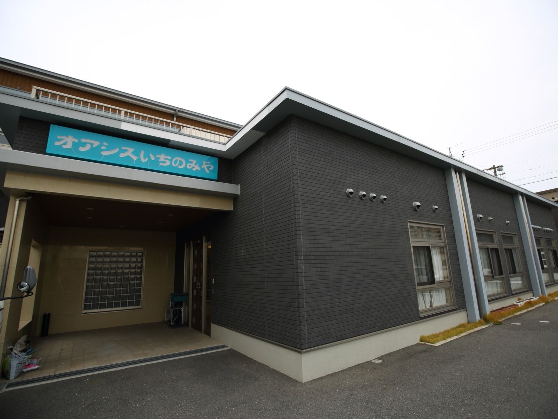 ダークグレーの外壁に明るいブルー系の施設プレートが印象的な施設外観である。平屋建てで、入口は奥まって設計されている。