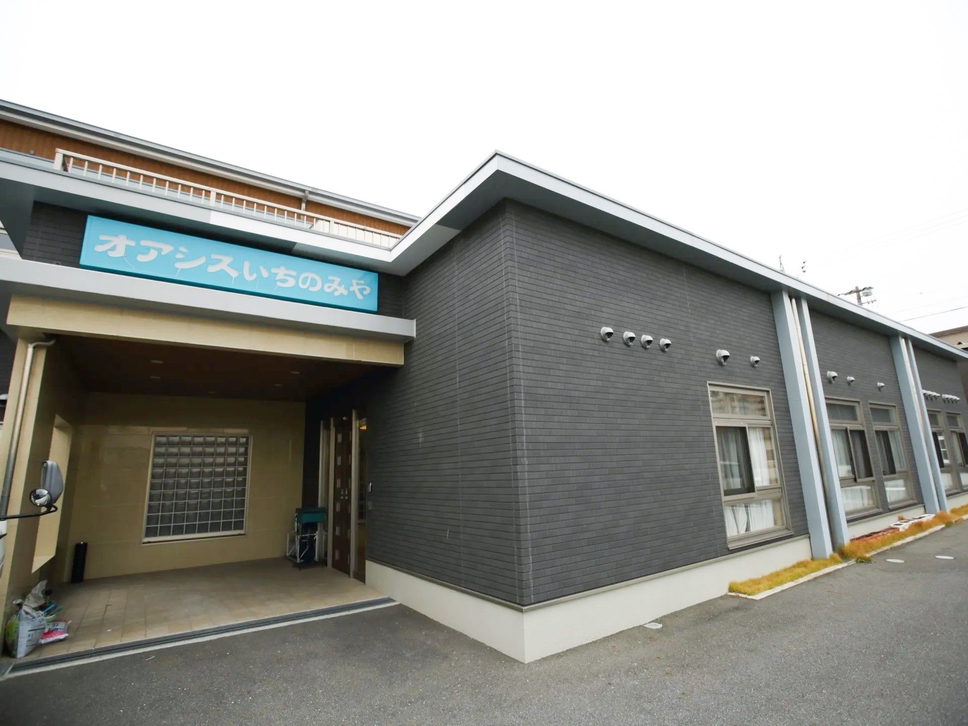 ダークグレーの外壁に明るいブルー系の施設プレートが印象的な施設外観である。平屋建てで、入口は奥まって設計されている。