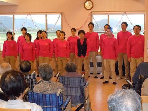 合唱している様子を写したもの。手前にそれを眺めている高齢者の様子