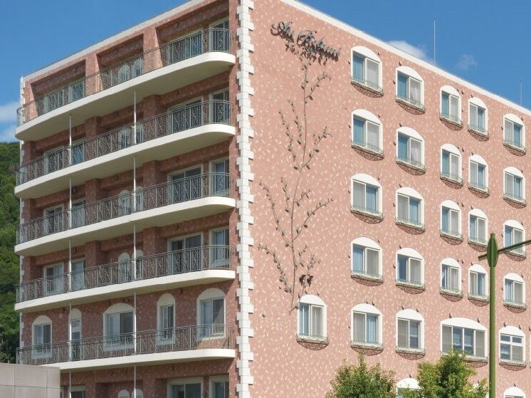 ブラウン系の建物は大きく、窓がたくさんあり、各フロアにベランダも付いていて、建物周辺には緑も多く植えられている。