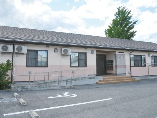 広い駐車場がある平屋建ての建物の玄関には手すり付きのスロープがあり、ポストも設置されている。