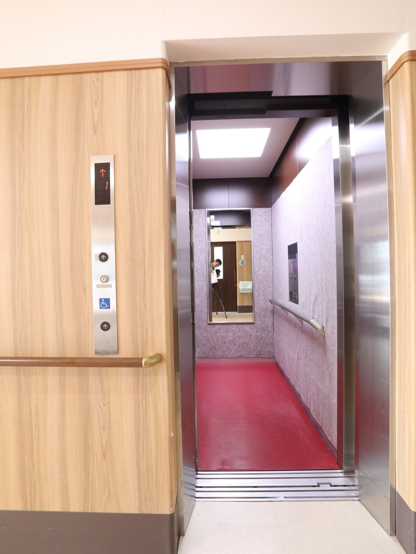 奥行に長いエレベーター内部である。正面に鏡があり、横側の壁に手すりを設置している。天井の照明は正方形である。