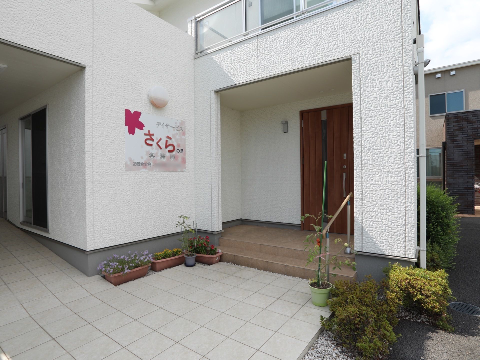 建物から奥に、木目調の玄関ドアがある。前方には、２段の階段があり、横側に手すりを設置している。両側に低木と花のプランターを飾っている。