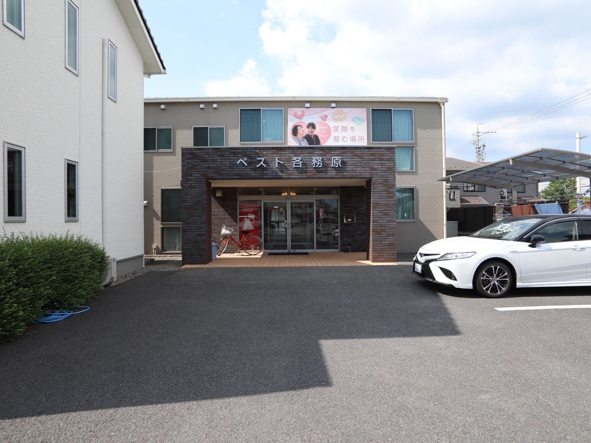 正面にグレーの２階建て、横側に白い外壁の建物がある。敷地内には、１台の車が停められている。周辺は閑静な住宅街である。