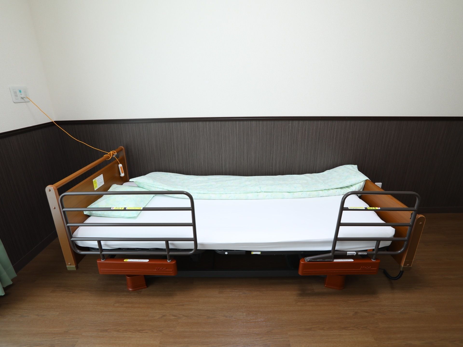 部屋の壁際に置かれたベッドである。ベッドガードが２ヵ所に設置されており、壁には、呼び出しボタンがかかっている。
