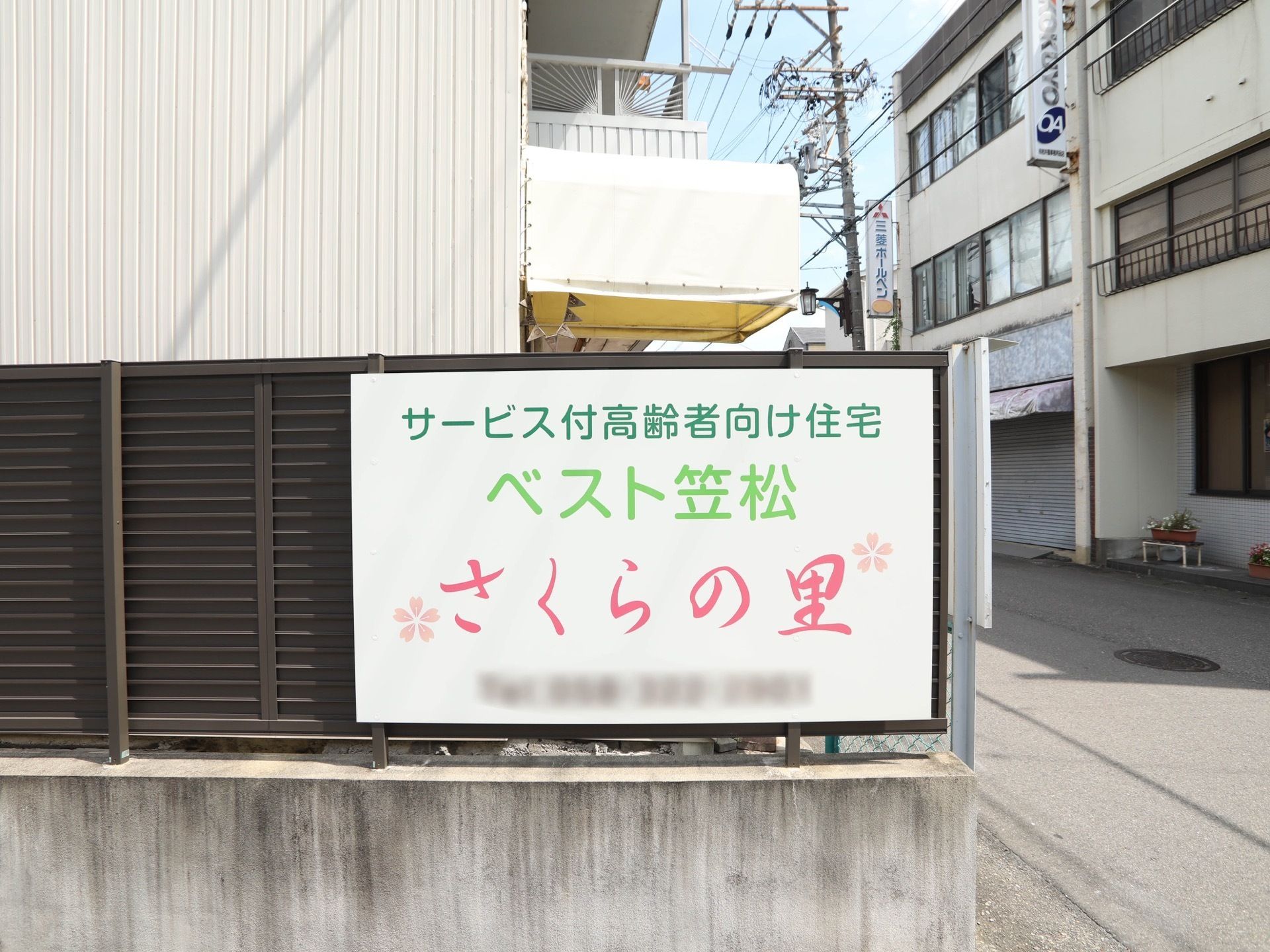 こげ茶色のフェンスに、施設名が書かれた看板がかかっている。周囲には、３階建ての建物が並んで建っている。