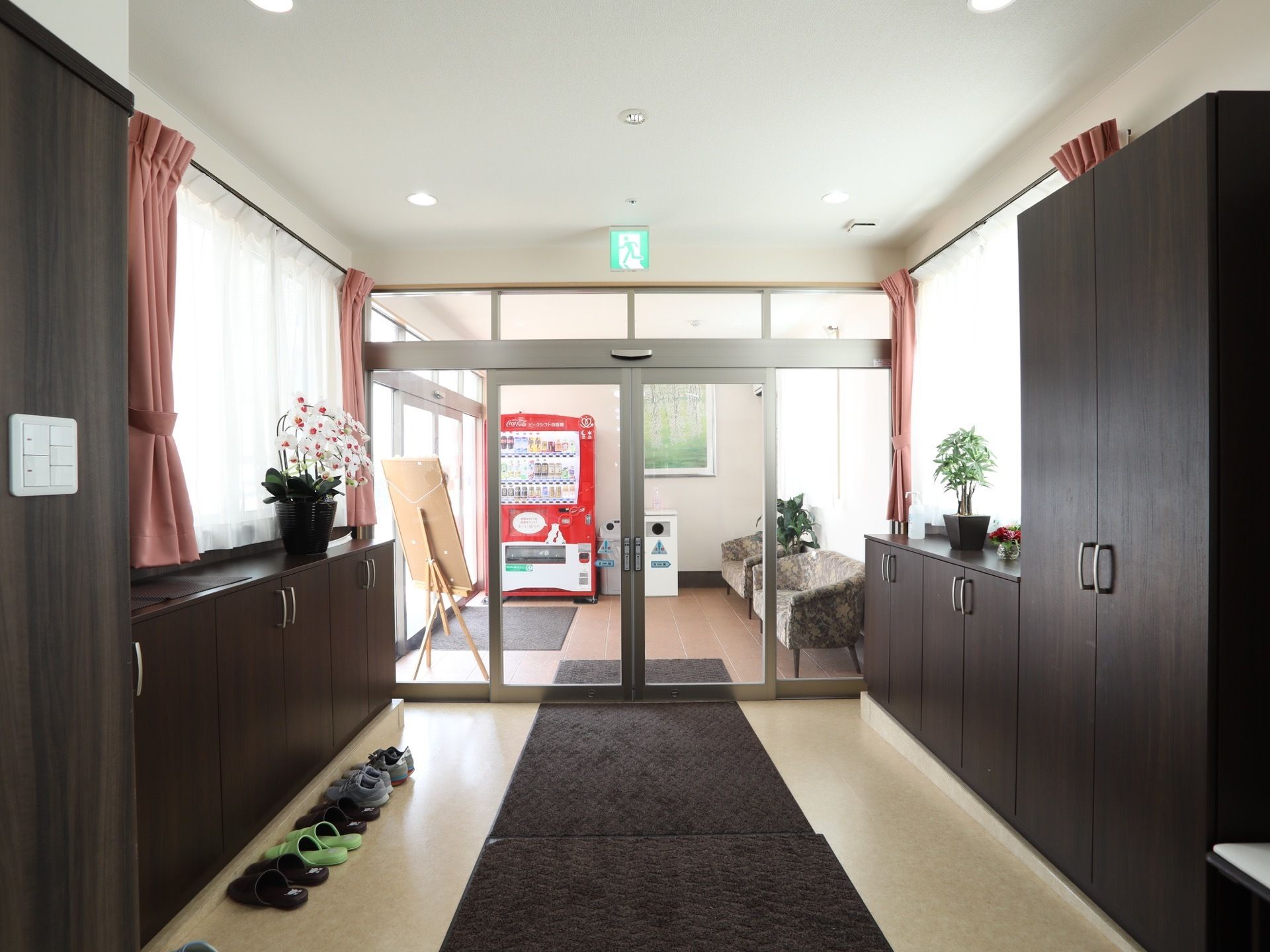 入口の両側に扉付きの棚がある。棚の上には鉢に入った白い花が飾られている。天井付近には非常口の案内灯がある。