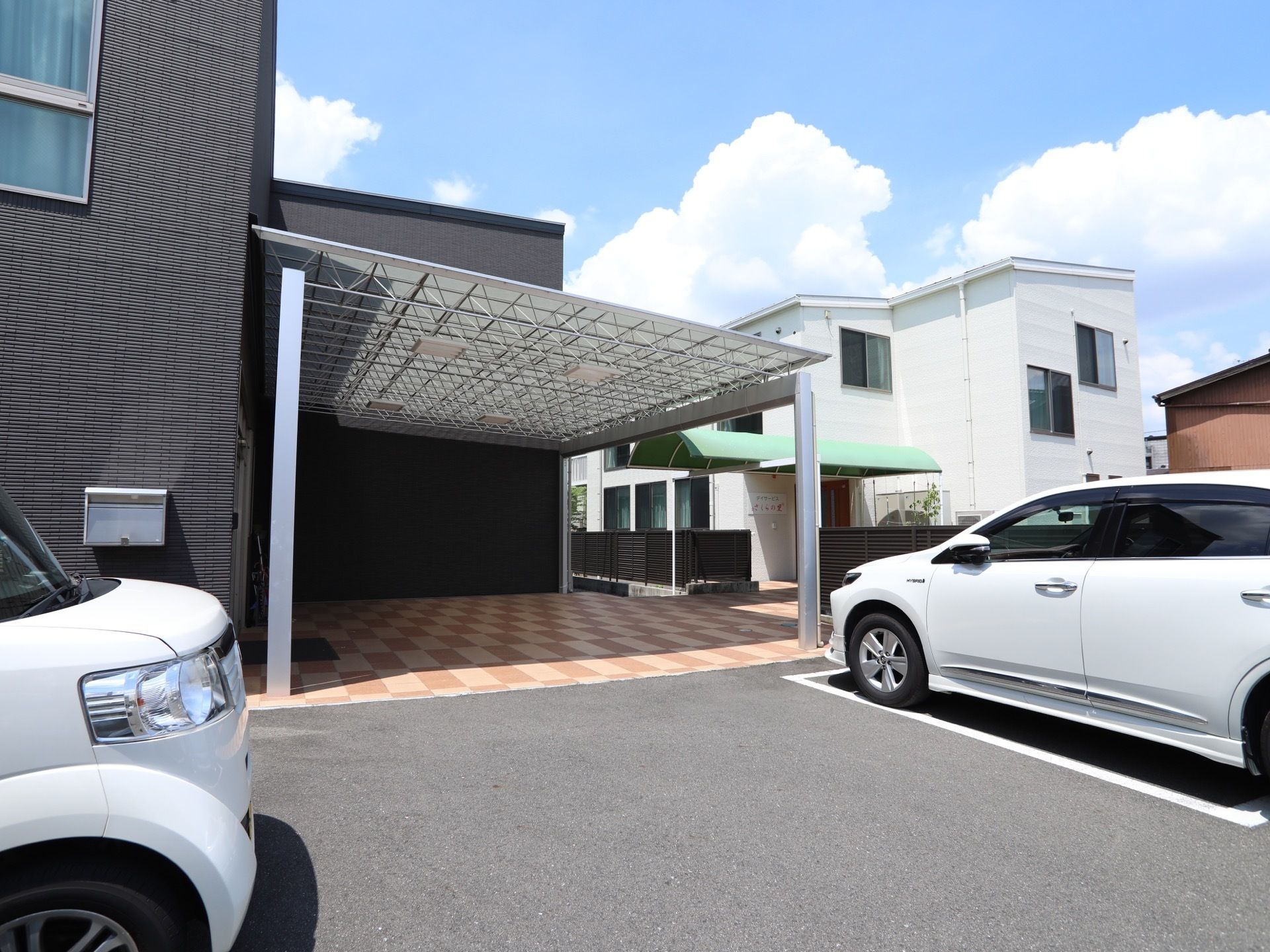 黒い外壁の建物に併設された、屋根付きのスペースがある。また、白色の車が２台、駐車場に停められている。