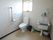 白い壁のトイレである。壁にはペーパーホルダーと手すりがあり、深い洗い場が併設されている。床には、掃除道具が置いてある。