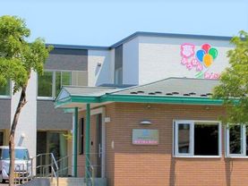2階建ての建物。モノトーンの建物の右手には茶色と白の外壁の併設事業所があり、玄関からは手すり付きのスロープが伸びている。
