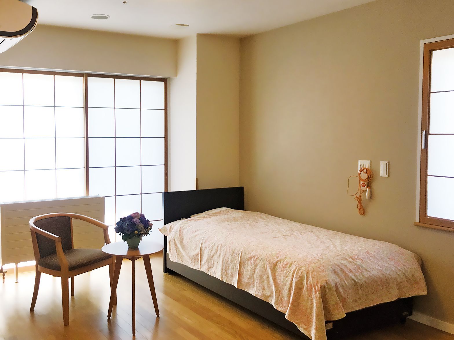 障子が設置された大きな窓があるフローリングの居室には、ベッドやテーブルセットがあり、壁には緊急通報装置が付いている。