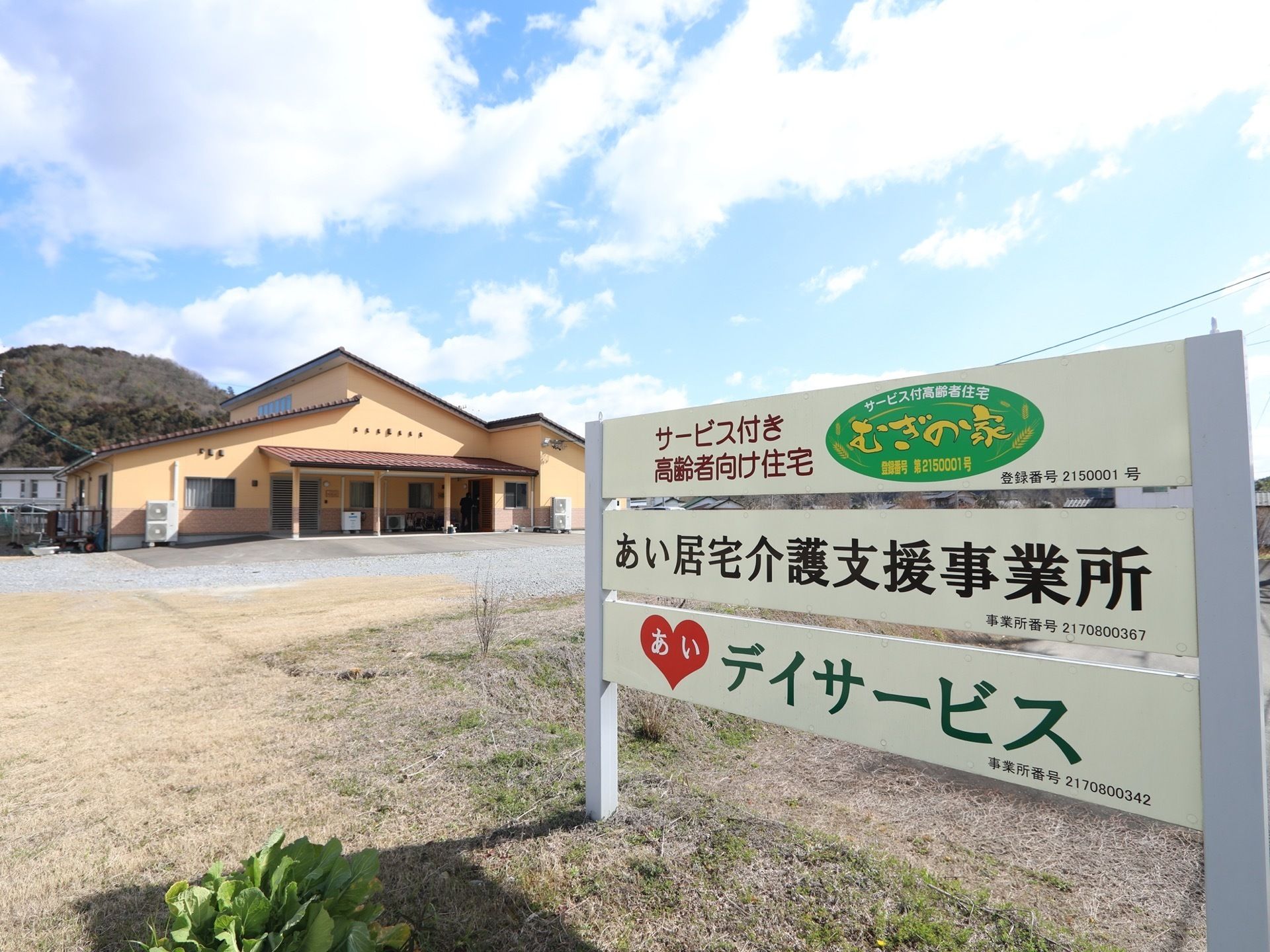 敷地の入口には施設名が書かれた白い看板が立てられている。奥の方には施設の建物があり、その前は広いスペースになっている。