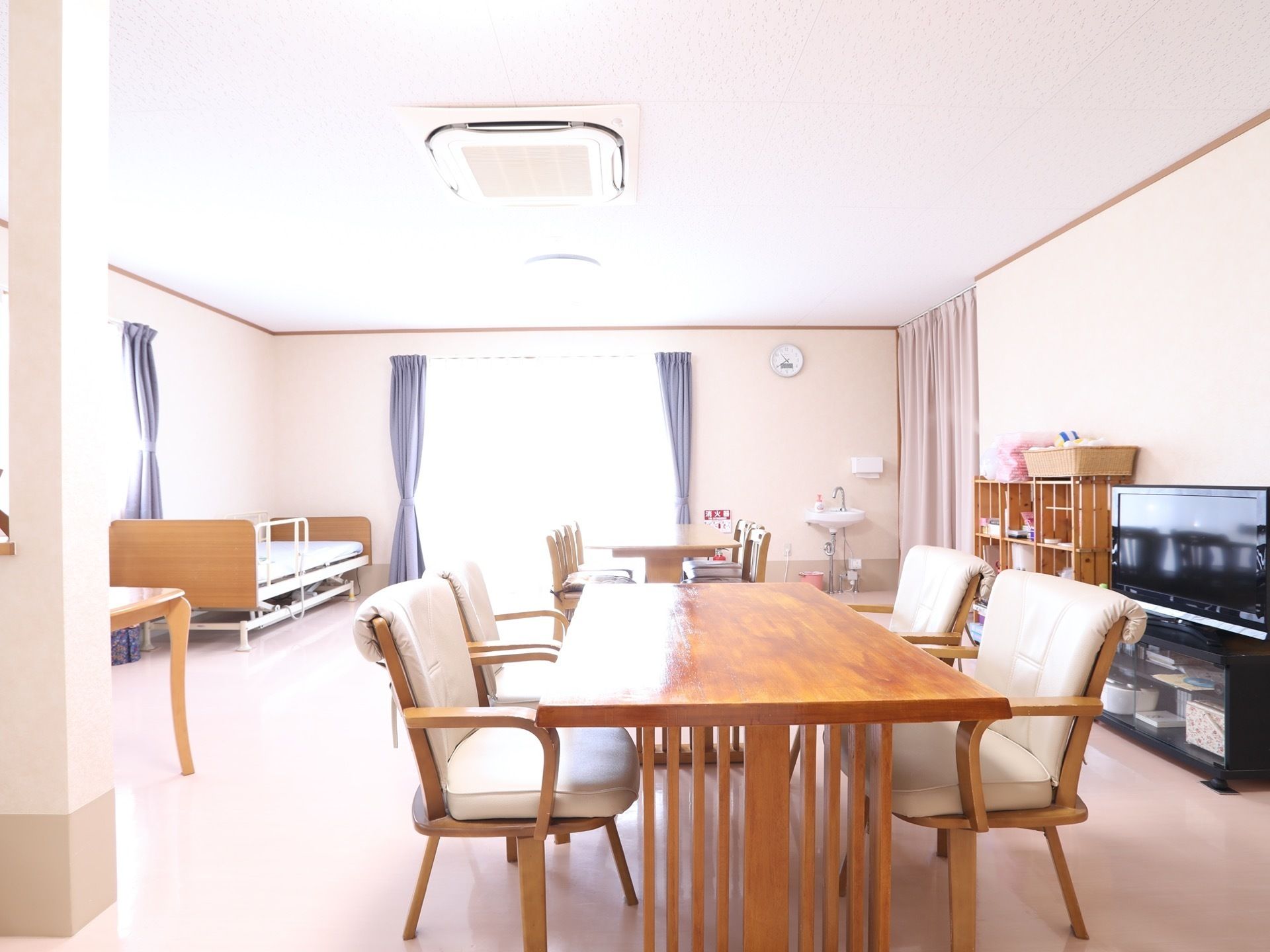 明るい食堂には木目調のテーブルと白い椅子がある。。テレビや洗面台が設置されており、奥の掃き出し窓にはカーテンが束ねられている。