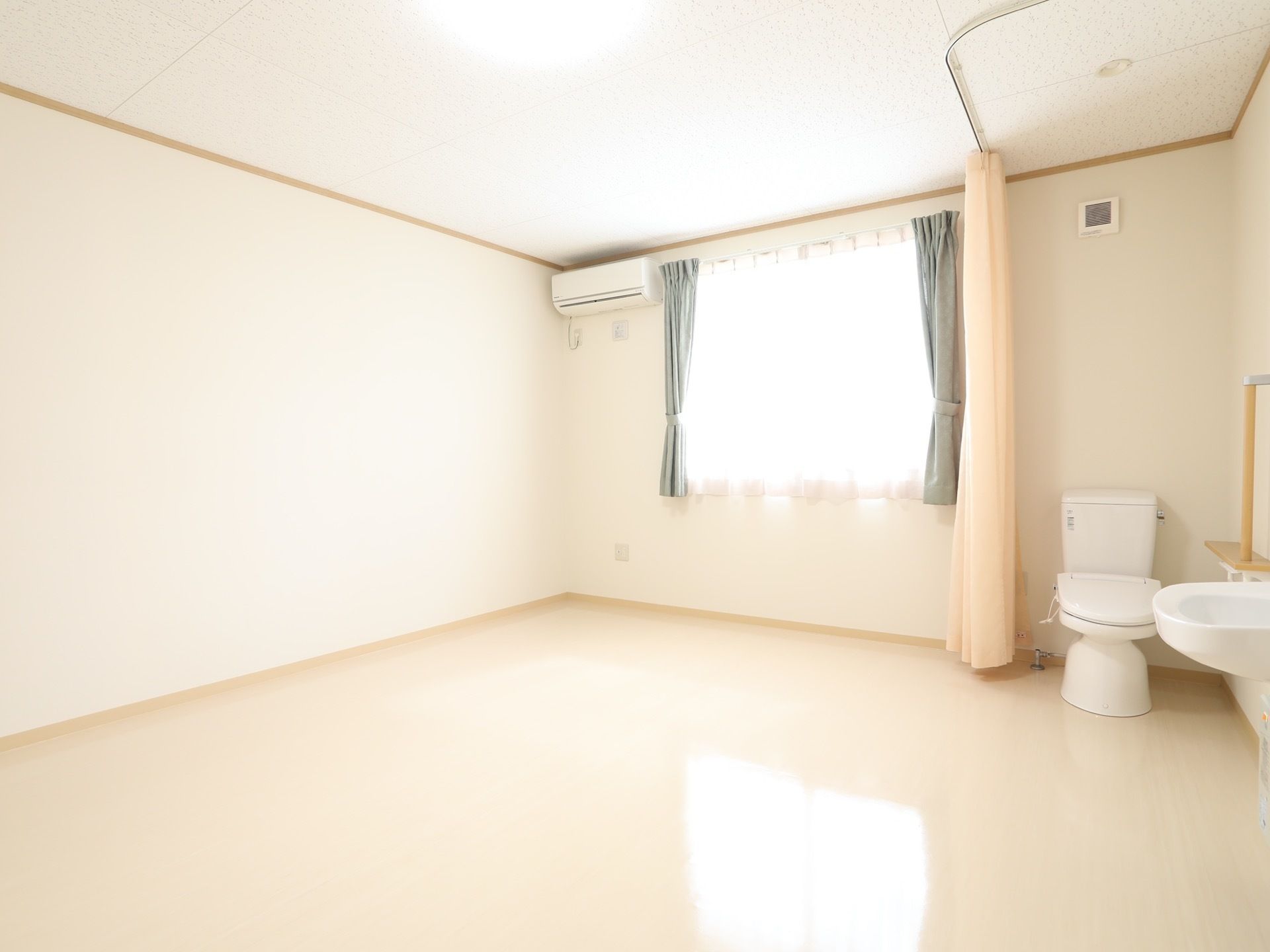 居室には腰高窓があり、レースカーテンとブルー系のカーテンが付けられている。天井近くの壁にはエアコンが設置されている。