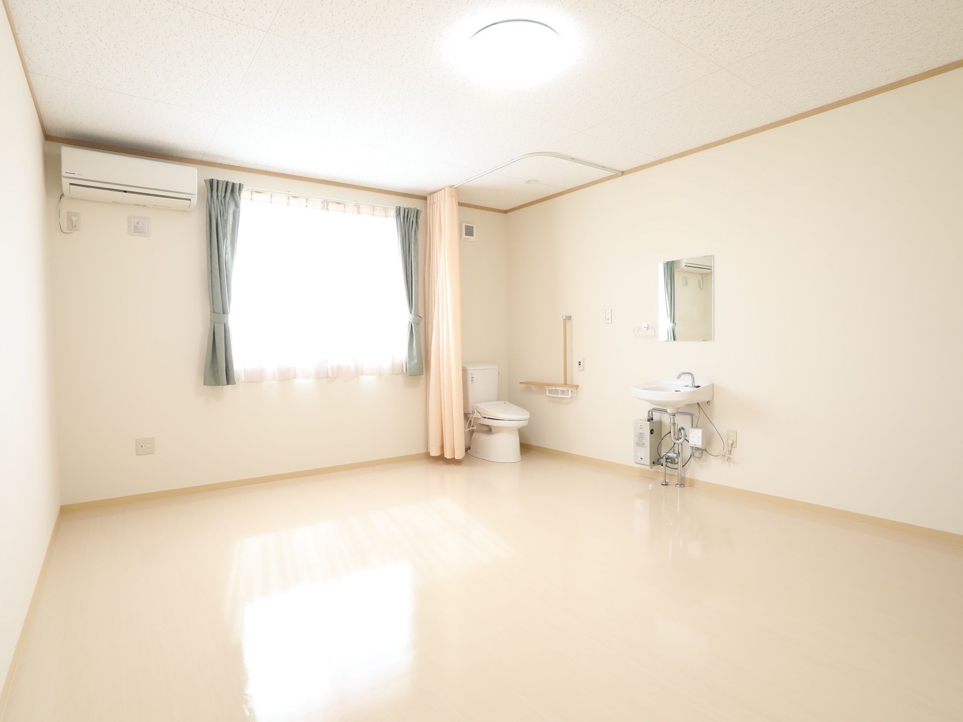 広い居室には洗面台とトイレが設置されている。トイレを囲むように白いカーテンが取り付けられていて、便器の横の壁にはL字型の手すりが備えてある。