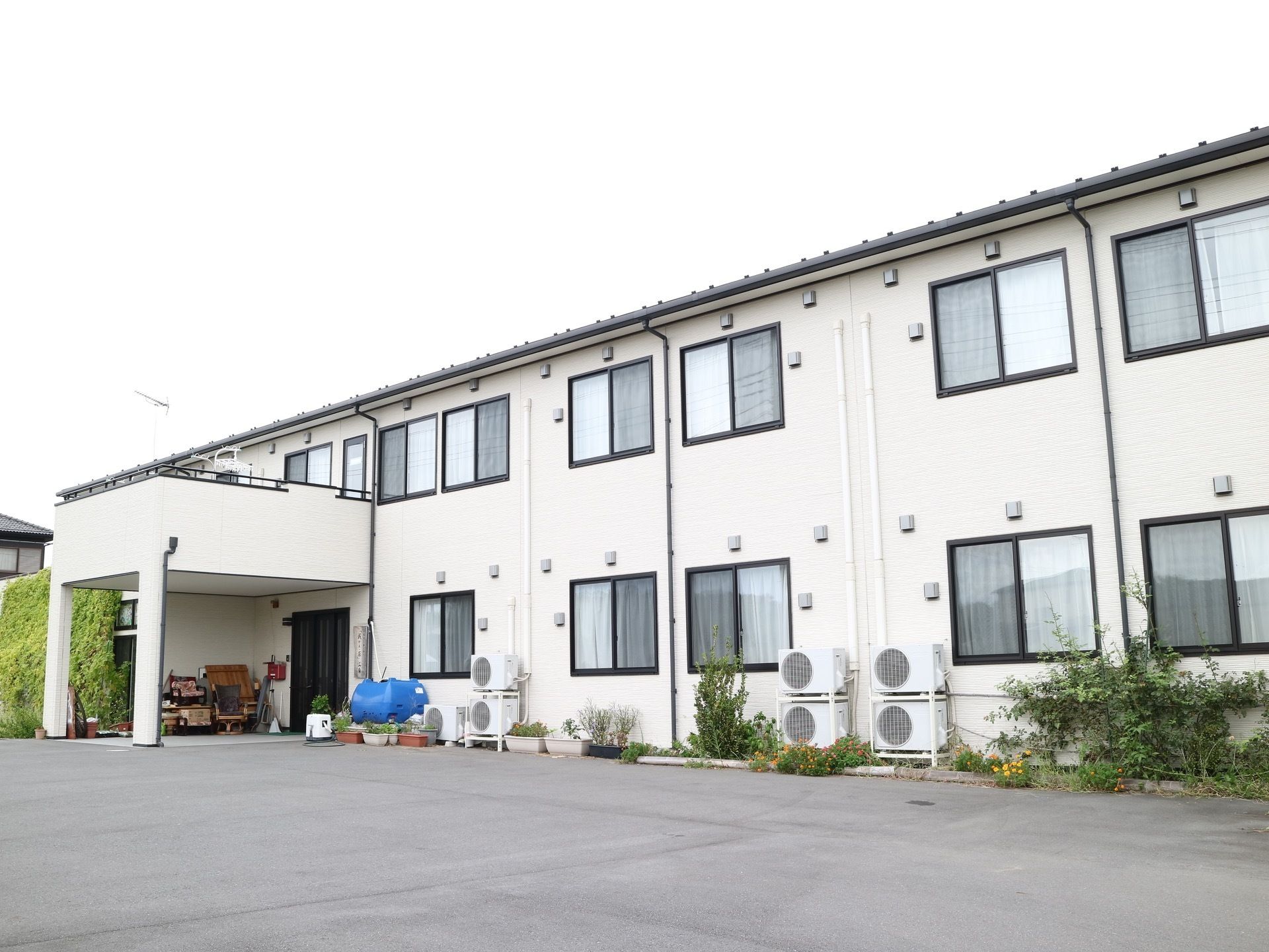 ２階建ての白い建物があり、１階部分にはエアコンの室外機が並んでいる。入り口付近にはプランターがいくつか置かれている。