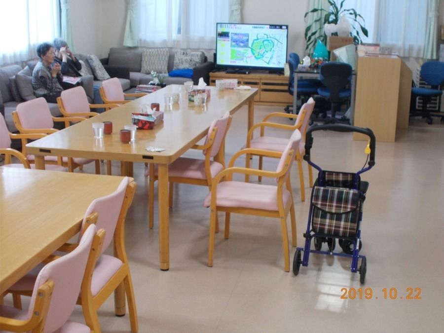 ピンクのエプロンを付けた女性スタッフが黄色いカーディガンを着て車いすに座っている高齢女性の方と手に自分の手を置き、微笑んでいる様子。