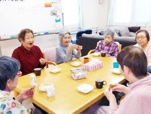 白いニット帽をかぶった車いすの高齢女性が外で一休みをしている様子。そばにはブルーのエプロンを付けた女性スタッフがいて、女性の手を取り微笑んでいる。