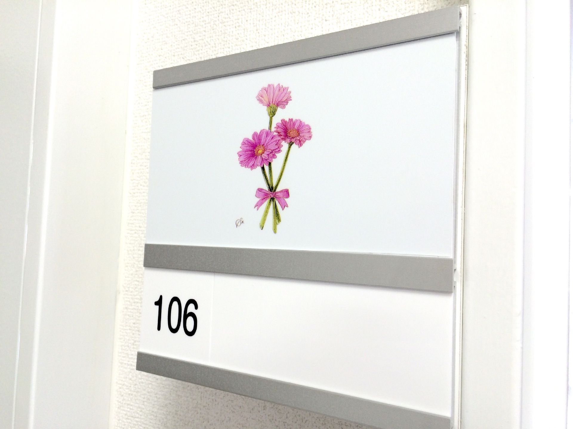 ピンクの花が3厘、ピンクのリボンでまとめられているイラストが描かれたネームプレート。部屋番号は「106」で、黒い文字で書かれている。