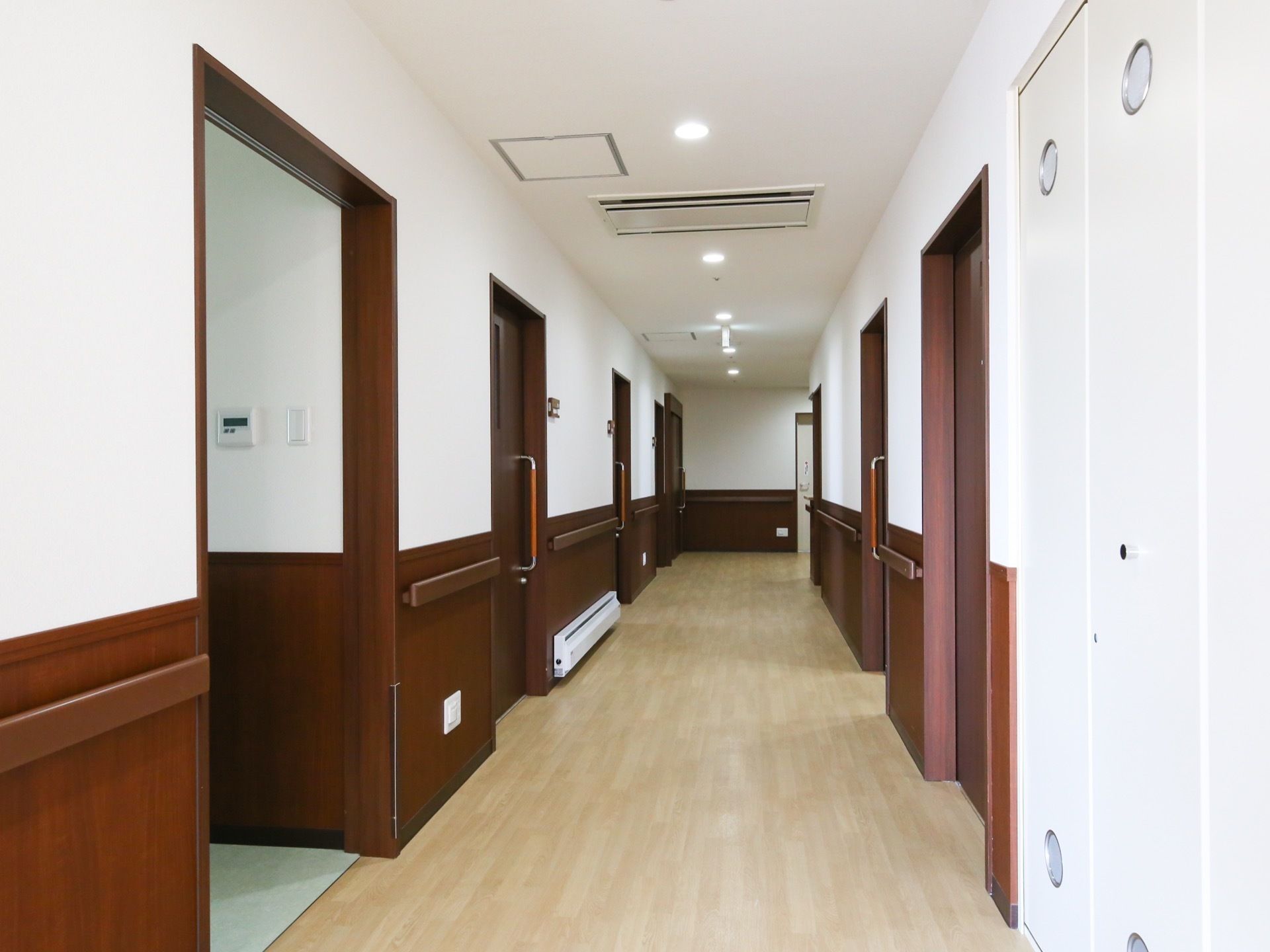 濃い茶系のスライド式居室扉が設置された廊下である。壁には、手すりや足元を温めるヒーターが完備されている。