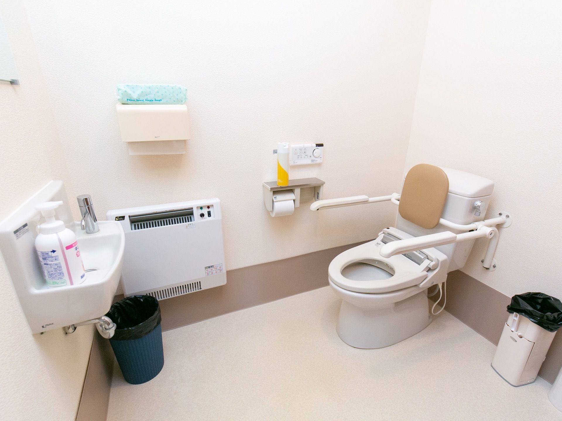 広々として掃除が行き届いたトイレである。便座には手すりと背もたれが設置されており、壁にはヒーターなどが取り付けられている。