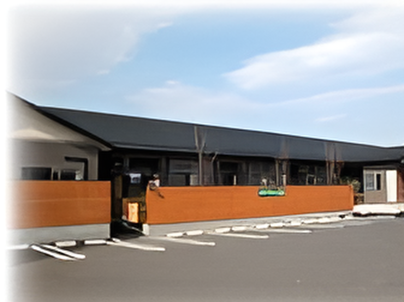 広い駐車場が付いた平屋建ての建物はグレーの三角屋根が付き、オレンジの外壁で日当たりのいい場所に建っている。