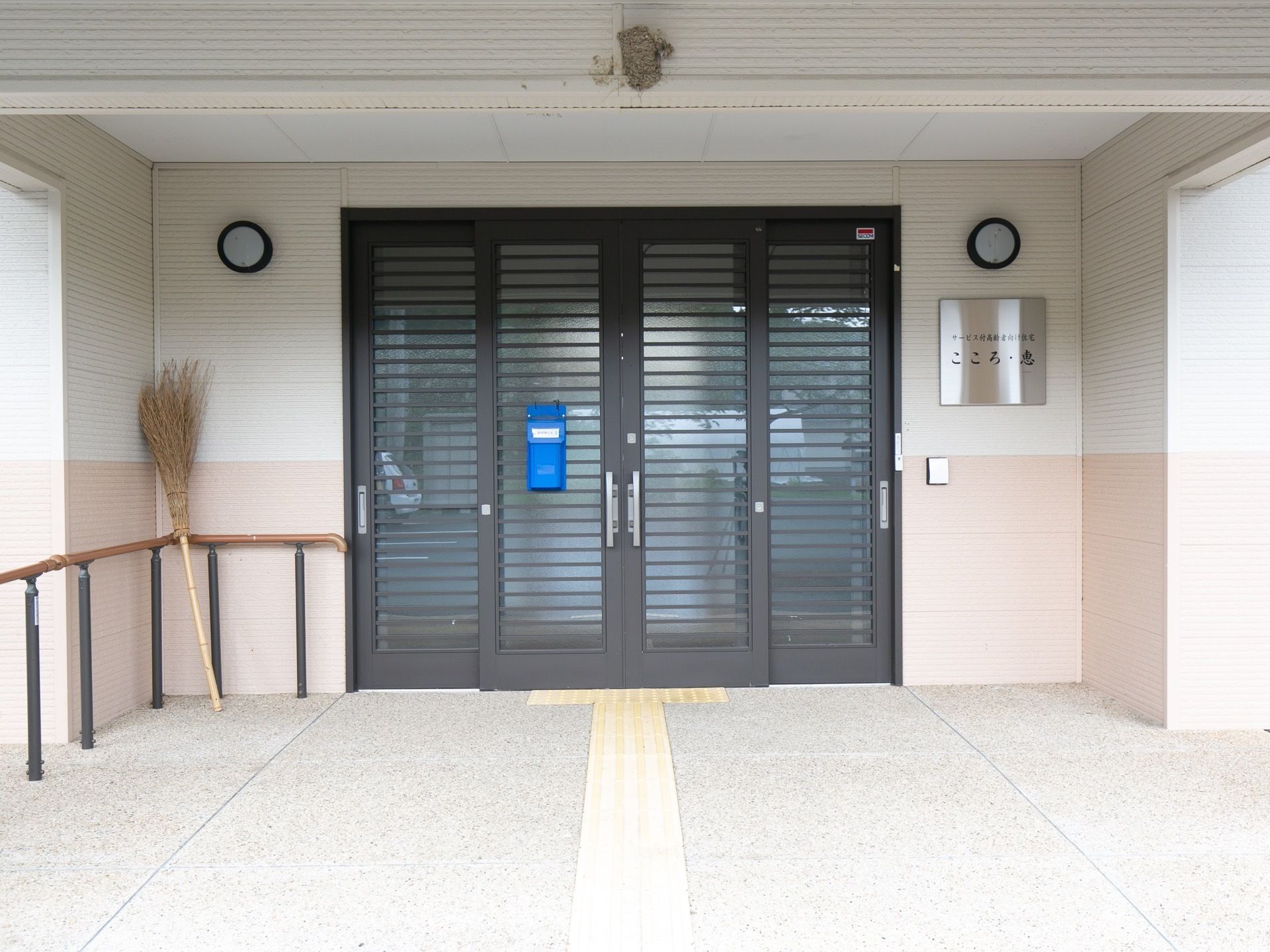 スライド式の扉が設置されている玄関で、床には黄色い点字ブロックがある。外壁には、表札や照明がかかっている。