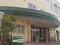 ベージュ系の建物は３階建てで、玄関は自動ドア。玄関脇には木もあり、車寄せもついている。