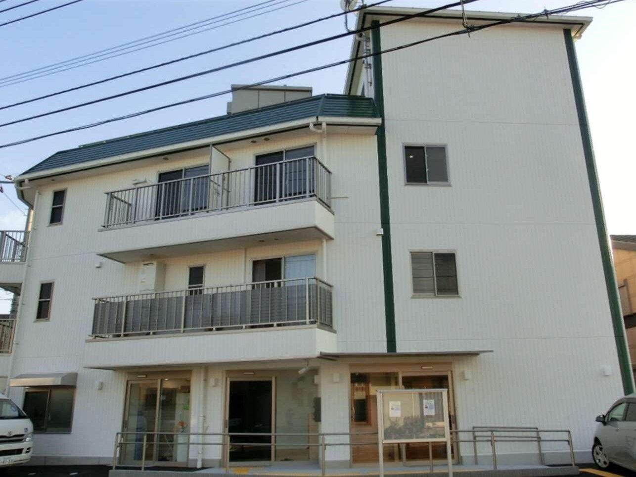 白い３階建ての建物。玄関に屋根と手すりが付き、各フロアにベランダがあり、駐車スペースには車が止まっている。