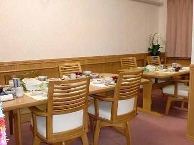 食事が並んだ木製のダイニングテーブルが二組並んだスペース。壁には手すりがあり、千羽鶴も飾られている。