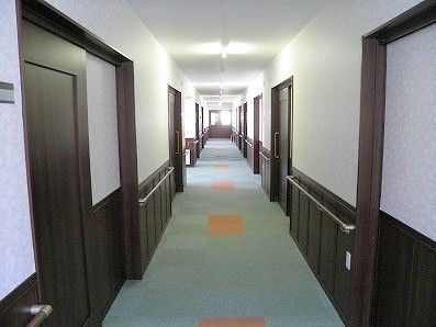 両サイドに手すりと居室のドアが並んだまっすぐな廊下。フロアにはグリーンのカーペットが敷かれ、居室の間はオレンジ色になっている。