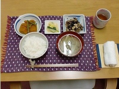 白いご飯にみそ汁、あえ物や煮物などがランチョンマットの上に並び、横には湯飲みに入ったお茶とお絞りがある。