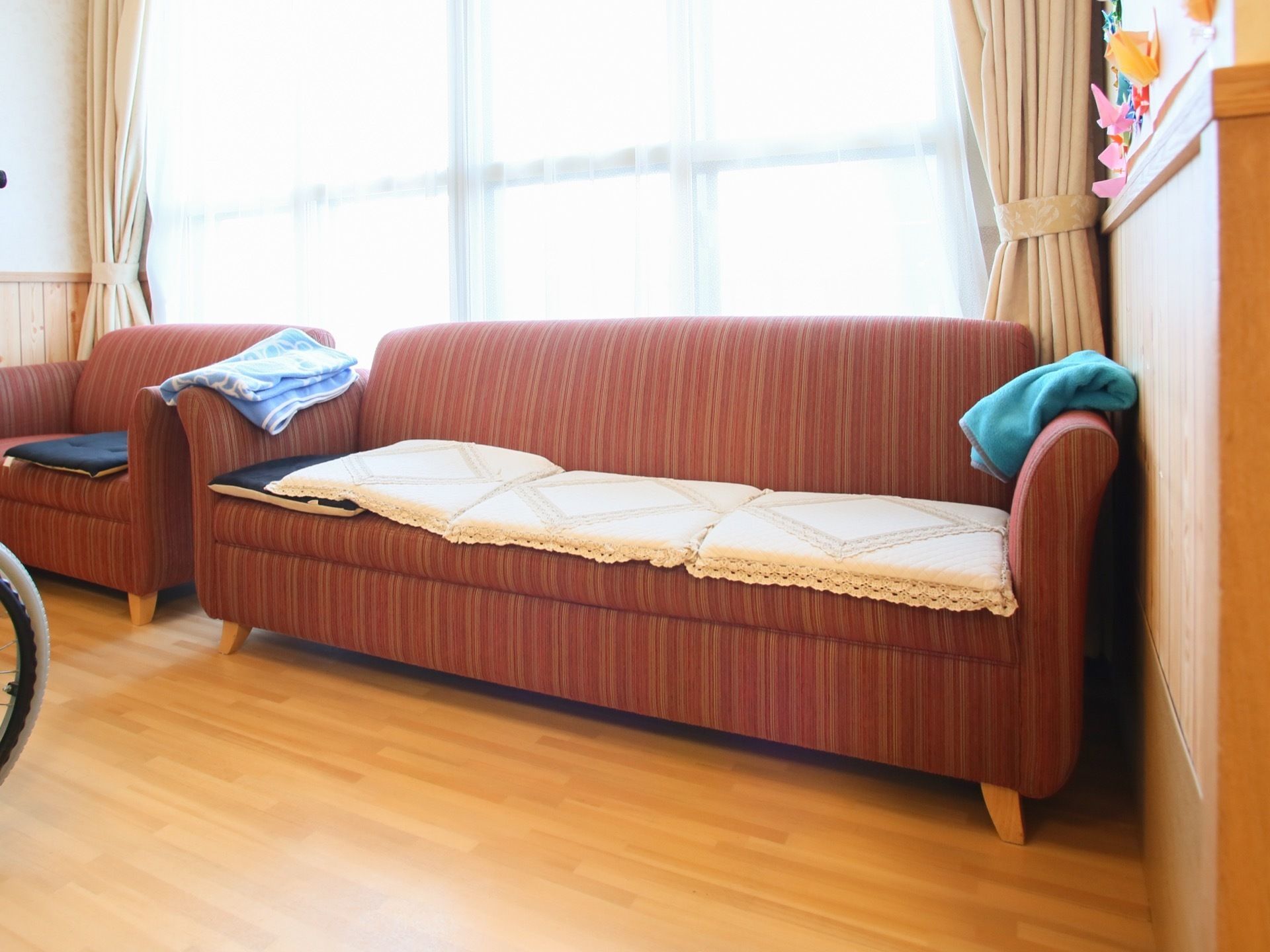窓のすぐ側に、一人用のソファと３人掛けのソファが並べて置いてある。ひじ掛けにはひざ掛けがあり、ソファの上にはクッションが敷いてある。