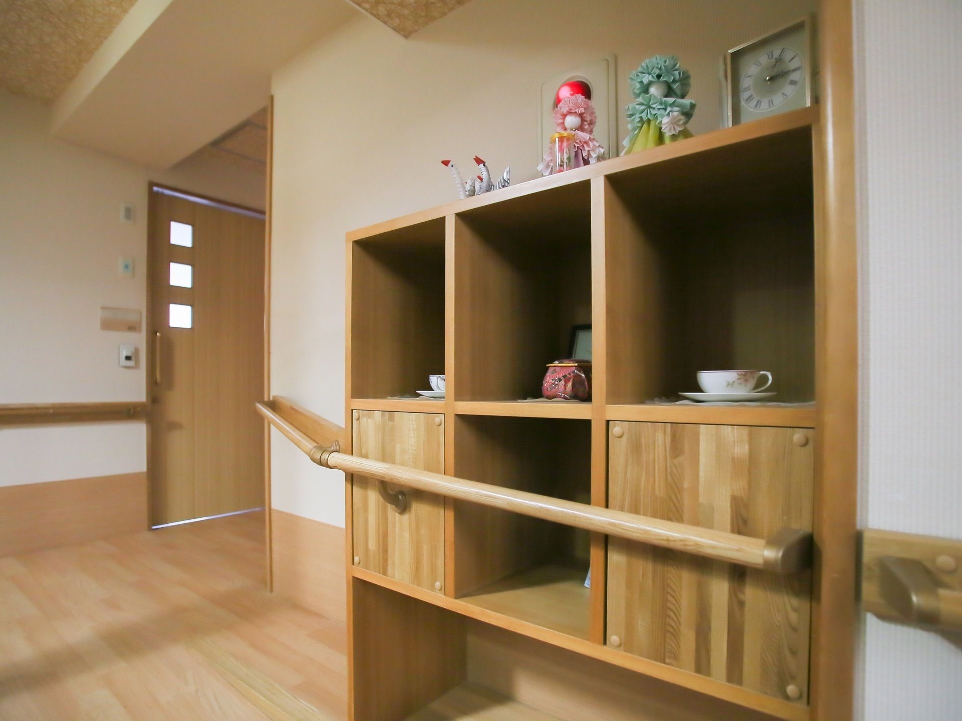 入居者様の居室のある廊下の一角。四角い飾り棚があり、時計や人形、コーヒーカップが飾られている。棚の中央付近には手すりが設置されている。