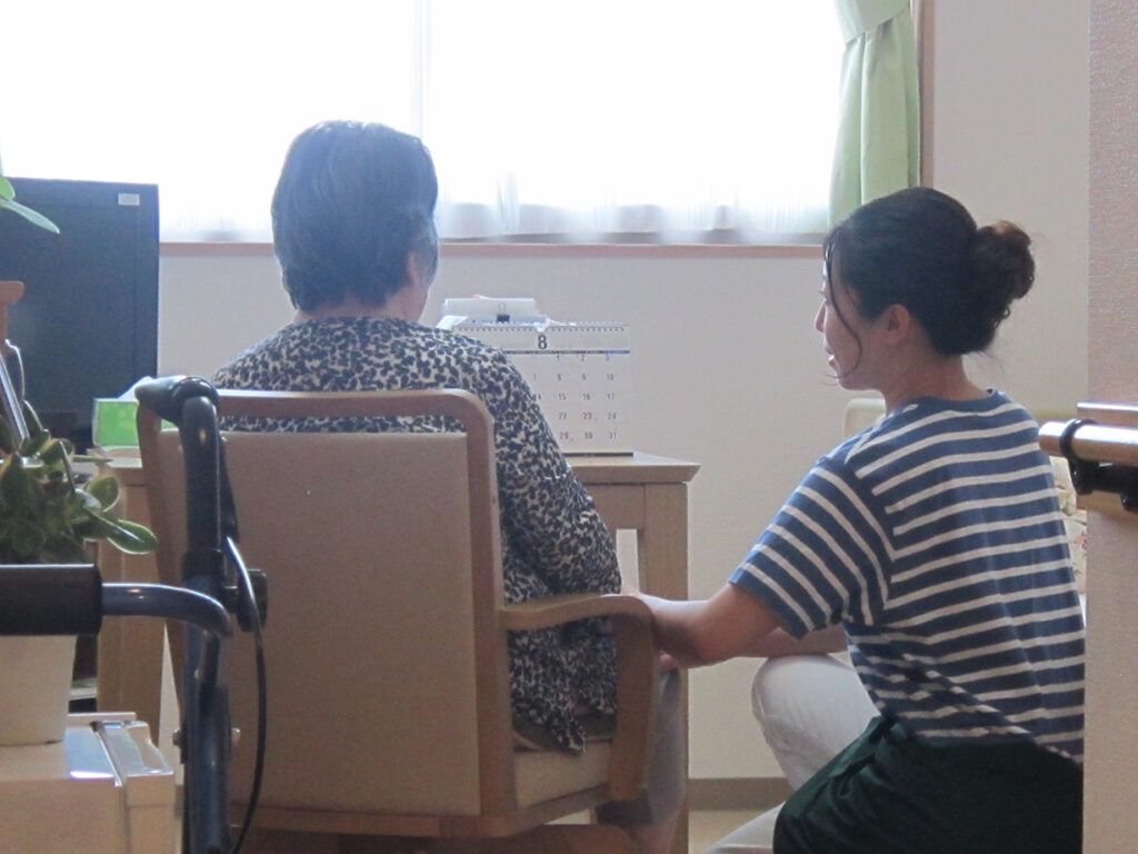 ボーダー柄の服を着た女性スタッフががいすに座った高齢女性に寄り添っている様子。