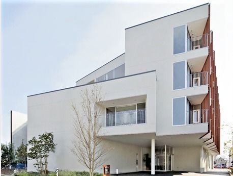 白を基調とした4階建ての建物。1階部分のエントランスが少し奥まったところにあり、上のフロア部分が屋根の役割をしている。