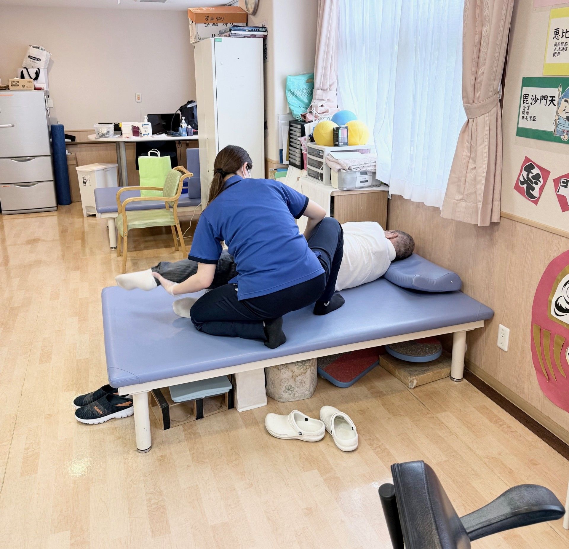 車いすの高齢女性は鶯色のカーディガンを着ていて、そのそばには紺のポロシャツを着た女性スタッフがいる。