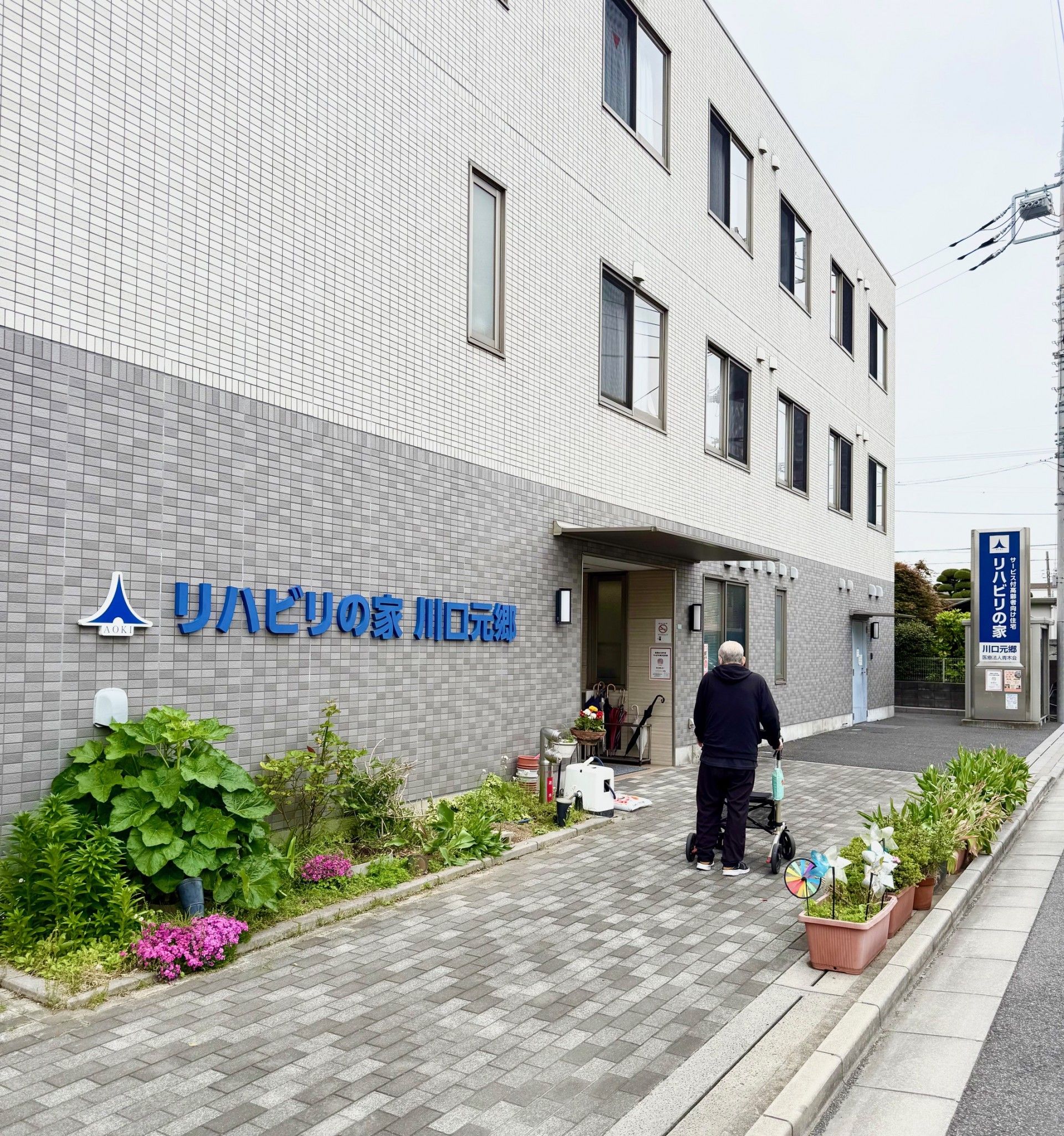 ピンクのエプロンを付けた女性スタッフが車いすの高齢女性と一緒に日当たりのいい屋外を散歩している様子。
