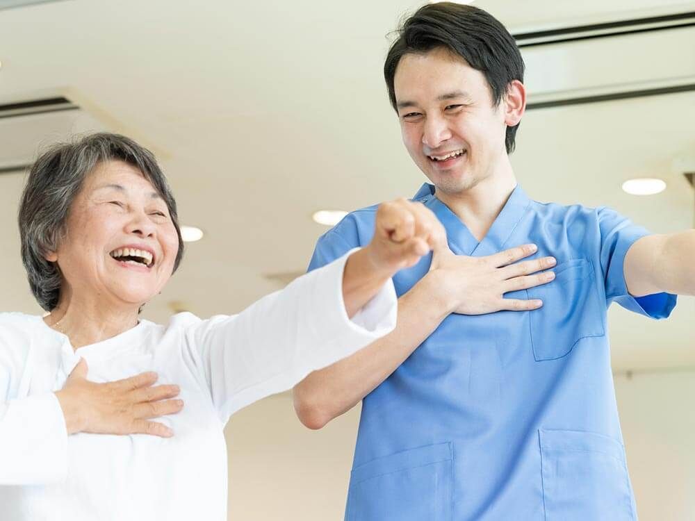 白いシャツを着た高齢女性とブルーのユニフォームを着た男性スタッフが笑顔で一緒に体操をしている。