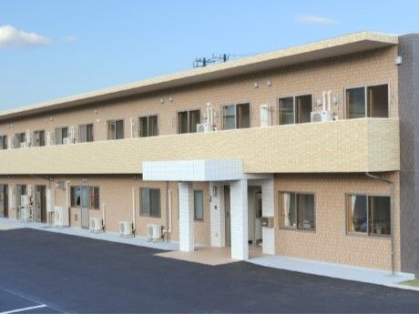 広い駐車場が付いた２階建ての建物は日当たりのいい場所に建っている。外壁はベージュで車寄せが付いている。